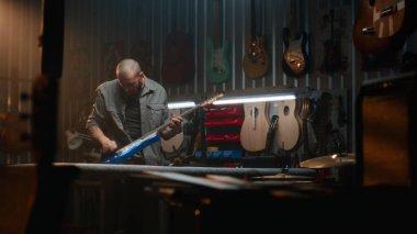 Erkek marangoz gitar tellerini bez parçasıyla siliyor ve gitarı inceliyor. Ahşap ustası, tasarımcı modern atölyede ahşaptan müzik aleti yapıyor. El işi ve küçük işletme konsepti.