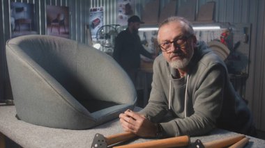 Profesyonel erkek marangoz sandalyeye yakın duruyor ve kameraya bakıyor. Olgun bir zanaatkar atölyede ya da stüdyoda montaj, üretim ya da yenileme mobilyaları için çalışır. El yapımı ve girişimcilikti. Portre.