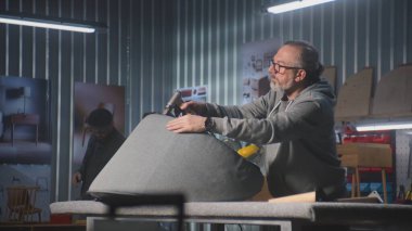 Olgun marangoz koltuğu zımba tabancasıyla döşenmiş. Erkek meslektaşım rafları arka planda monte ediyor. Sanatçılar mobilya yapmak veya restore etmek için şık bir atölyede çalışırlar. El işi ve küçük işletmeler..