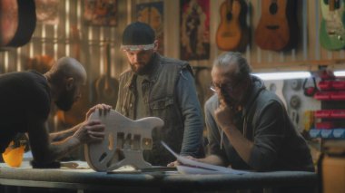 Olgun beyaz marangoz planlara bakar ve şık bir ışık atölyesinde çalışmayı tartışır. Erkek dövmeli zanaatkar gitar gövdesini tutuyor ve iş arkadaşlarıyla konuşuyor. El yapımı ve girişimcilik kavramı.