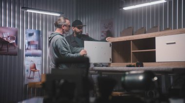 Profesyonel erkek marangoz ya da tasarımcı dijital tablet bilgisayar kullanır, olgun müşterilerle konuşur ve ahşap TV standı yapmayı tartışır. Modern mobilya atölyesi. El yapımı ve girişimcilik kavramı.