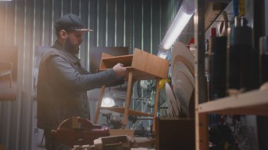 Profesyonel marangoz, profesyonel aletlerle LED lambanın altında ahşap komodin monte eder. Erkek zanaatkar, esnaf mobilya yapmak için modern atölyede çalışıyor. El işi ve küçük işletme konsepti.