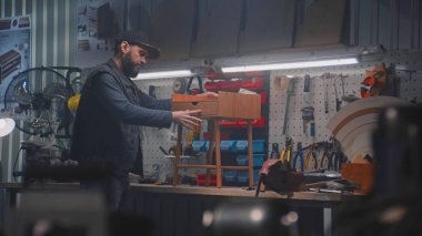 Erkek marangoz modern mobilya atölyesinde çalışan ahşap komodinleri yapar veya monte eder. Başka bir zanaatkar, zanaatkar aletlerle birlikte gemide asılı duruyor. El yapımı ve girişimcilik kavramı.
