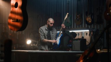 Erkek zanaatkar, kombinasyon amfisine bağlı gitar çalar, sesi kontrol eder. Marangoz modern atölyede ahşap müzik aleti yapıyor. Arka planda başka bir zanaatkar çalışıyor. El yapımı ve girişimcilik..