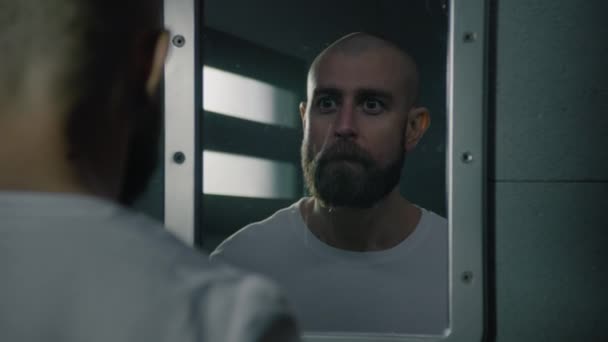 沮丧和沉思的男性囚犯站在那里 在监牢的镜子里看着自己 被非法定罪的人在狱中服刑 拘留中心或教养所 — 图库视频影像
