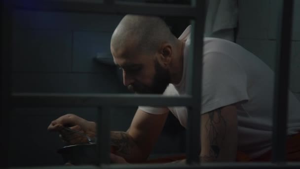 身穿橙色制服的纹身囚犯坐在牢房的床上 吃铁碗里的监狱食物 男囚犯因在狱中犯罪而服刑 拘留中心或教养所 — 图库视频影像