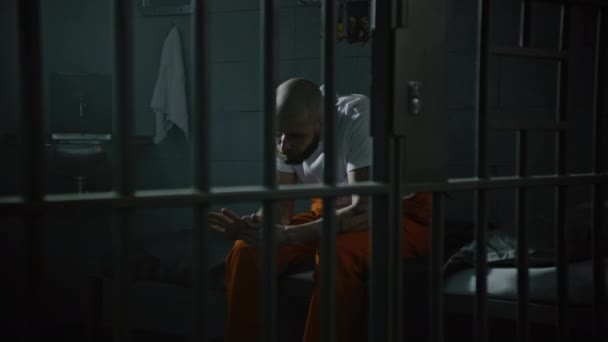 身穿橙色制服的罪犯坐在牢房的床上 犯人因在狱中犯罪而服刑 Gangster在拘留中心教养所司法系统 透过金属棒观看 — 图库视频影像