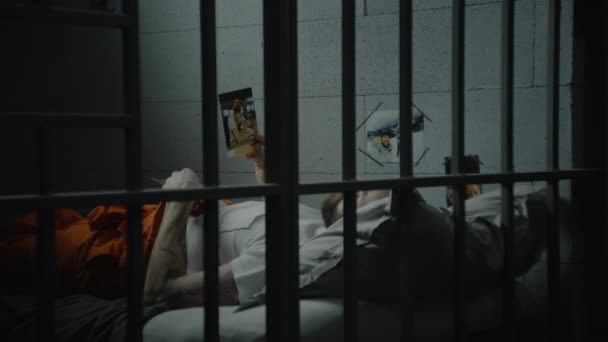 男囚犯躺在牢房的床上 看着家人的照片 犯人或犯人因在狱中犯罪而服刑 通过金属棒观看 拘留中心或教养所 — 图库视频影像