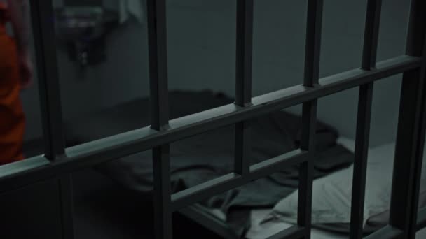 オレンジ色の制服の犯罪者は刑務所のベッドに横たわっている 刑務官は犯罪の刑期を定めている 拘置所で同行する 矯正施設 司法制度 金属棒を通して見る クローズアップ — ストック動画
