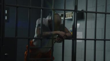 Turuncu üniformalı depresyondaki erkek mahkum hücrede duruyor metal parmaklıklara yaslanıyor ve el ele tutuşuyor. Mahkûm hapiste suçtan hapis yatıyor. Gözaltı merkezi ya da ıslah evi.