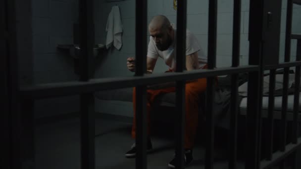 オレンジ色の制服の男性捕虜は刑務所のベッドに座り 鉄のボウルから嫌な刑務所の食べ物を食べようとします 刑務所では刑務所に収監されている 拘禁センターまたは矯正施設 — ストック動画