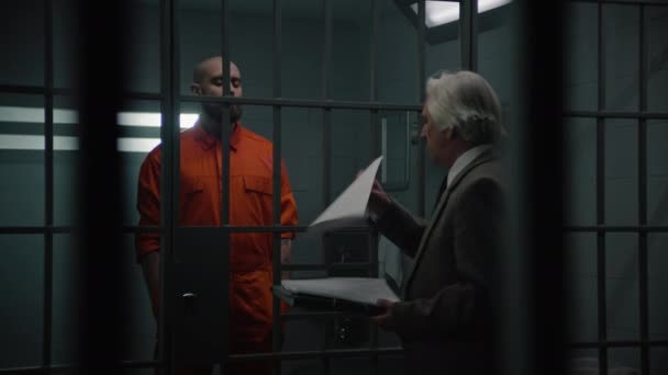 オレンジ色の制服の囚人は 刑務所の刑務所のバーの後ろに立って 弁護士協定を読みます インメイトは刑務所で有罪判決を受けた 拘禁センターまたは矯正施設 — ストック動画