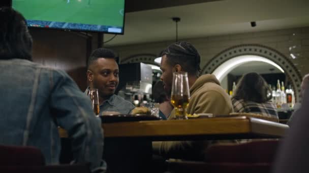 アフリカ系アメリカ人は夕方にスタイリッシュなバーに座って話す 男性の友人 スポーツファンは夕食を食べ 週末はスポーツバーで休む バックグラウンドでのライブフットボール試合のテレビ画面 — ストック動画