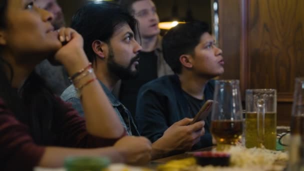 ヒンドゥーの男性は 夜に多文化の友人とパブのバーカウンターで座っているライブスポーツゲームを見ています ハッピースポーツファンは喜び サッカーの試合に賭け 携帯電話を使用してブックメーカーの評価をチェックします — ストック動画
