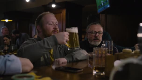 两个男性朋友坐在吧台边聊天喝啤酒 体育爱好者周末晚上在拥挤的酒吧里度过 电视屏幕上直播足球比赛 生活方式和休闲的概念 — 图库视频影像