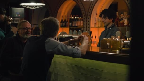 年轻的酒保把啤酒倒入杯子里 送给成熟的男人 两个男性朋友喝啤酒 晚上在酒吧柜台边与酒保交谈 生活方式和休闲的概念 — 图库视频影像