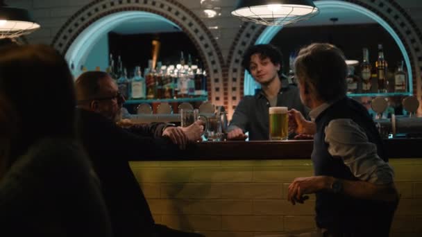 年轻的酒保给成熟的男性朋友们一杯啤酒 晚上两个男人坐在酒吧柜台边 在时尚的酒吧里聊天喝酒 遇见老朋友了 生活方式和休闲的概念 — 图库视频影像