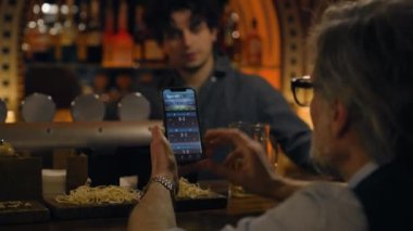 Olgun spor hayranları maç skorlarını kontrol ediyor ve uygulamada bahisçi reytinglerini kontrol ediyor, cep telefonu kullanıyor, barmenle konuşuyor. Erkek arkadaşlar bardaki tezgahta oturarak online futbol maçına bahis oynuyorlar. Kumar konsepti.