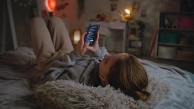Kulaklıklı genç bir kız evdeki yatakta uzanıyor, müzik dinliyor ve uygulamadan cep telefonu kullanarak şarkı seçiyor. Beyaz tenli genç, boş zamanlarını gündüzleri rahat yatak odasında geçirir. Yaşam tarzı kavramı.