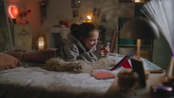 小女孩躺在舒适的床上 晚上休息 白人青少年与朋友聊天 并用手机上网 微笑着 孩子们在家里舒适的大卧室里呆着 — 图库视频影像