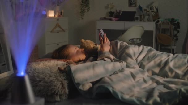 아름다운 소녀는 장난감과 침대에 그녀의 귀여운 여학생 저녁에 휴대폰을 인터넷을 — 비디오