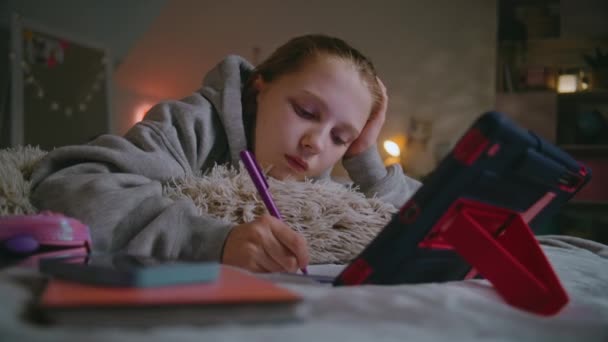 若い女の子は快適な寝室のベッドに横たわり ノートブックに書き込み タブレットコンピュータを使用して学校のレッスンを行います コーカサス人のティーンエイジャーは 昼間の自宅でオンラインで時間を費やし 勉強しています ライフスタイルコンセプト — ストック動画