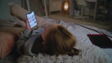 Kablosuz kulaklıklı genç beyaz kız evdeki yatakta uzanıyor, müzik dinliyor, sosyal ağları cep telefonu kullanarak kaydırıyor. Gençler boş zamanlarını rahat yatak odasında geçirirler. Yaşam tarzı kavramı.