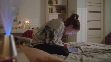 Genç kız odasında yatağında oturuyor ve internette geziniyor ya da tablet kullanarak internette sohbet ediyor. Hoş bir kız öğrenci boş zamanlarını evde sosyal ağları karıştırarak geçiriyor. Konforlu ve şık iç tasarımı olan bir ev..
