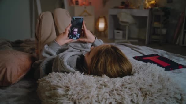 年轻的白种人女孩戴着耳机 用手机通过视频电话与朋友聊天 躺在舒适的卧室里 少女白天在家里消磨时光 生活方式的概念 — 图库视频影像