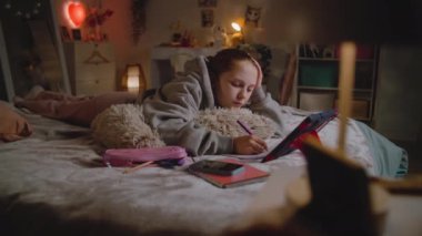 Genç kız evde yatakta yatıyor, deftere yazıyor ve dijital tablet bilgisayar kullanarak okul için online ödev yapıyor. Genç beyaz kız gündüzleri rahat yatak odasında vakit geçirir. Yaşam tarzı kavramı.