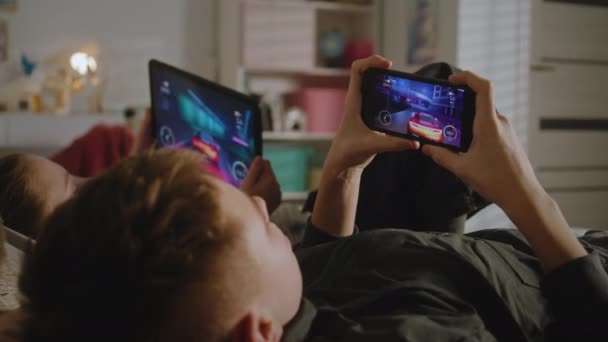 青少年躺在自己房间的床上 在数字设备上玩电子游戏 女孩用平板电脑 男孩用手机一起玩 姐姐和她哥哥在一起消磨时光 有舒适室内环境的家 — 图库视频影像