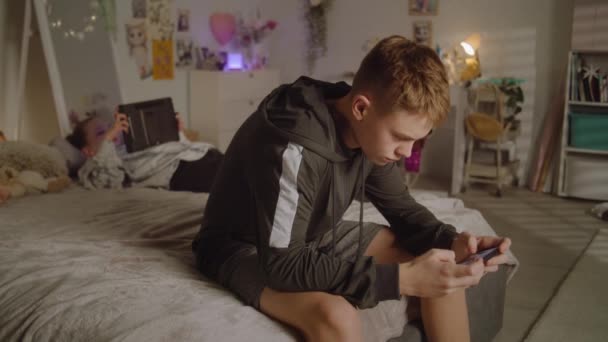 小女孩躺在床上 用平板电脑滚动社交网络 手里拿着手机的男孩在玩游戏 青少年的闲暇时间 家庭关系 时尚的家 — 图库视频影像