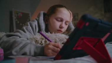 Genç beyaz kız evde yatakta uzanıyor, deftere yazıyor ve dijital tablet kullanarak online okul dersleri alıyor. Genç kız gündüzleri rahat bir yatak odasında ders çalışır ve zaman geçirir. Yaşam tarzı kavramı.