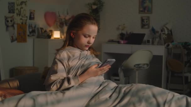 コーカサス人のティーンエイジャーは 朝早くから快適な寝室で起きる 若い女の子は携帯電話を取り出し 警報を鳴らした ティーンは居心地の良いベッドに座って ソーシャルネットワークでチャットするために彼女のスマートフォンを使用します — ストック動画