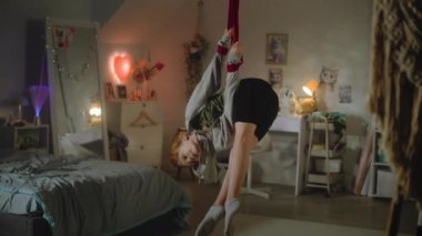 Beyaz tenli genç, açık yatak odasının ortasında akrobatik egzersizler yapmak için havadan jimnastik kurdeleler kullanıyor. Genç kız, büyük bir konfor odasında jimnastik yapıyor. Gençler evlerinde vakit geçirir..
