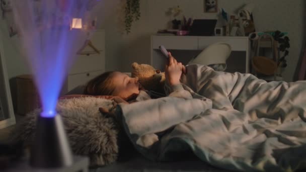 소녀는 장난감과 침대에 그녀의 아름다운 여학생은 저녁에 전화를 사용하여 온라인으로 — 비디오