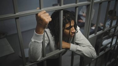 Kızgın Afrikalı Amerikalı ergen mahkum hapisteki hücrede duruyor, metal parmaklıkları var. Genç mahkumlar arka planda yatakta kağıt oynuyorlar. Gençlik gözaltı merkezi ya da ıslah evi. Yüksek açı.