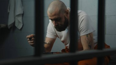 Turuncu üniformalı bir mahkum hücrede yatakta oturup demir kaseden hapishane yemeği yiyor. Erkek mahkum, hapishanede suçtan hapis yatıyor. Gözaltı merkezi ya da ıslah evi.