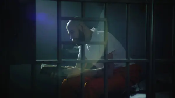 阅读圣经时 身穿橙色制服的男性囚犯坐在狱中的床上 被非法定罪的人在狱中服刑 通过金属棒观看 拘留中心或教养所 — 图库照片