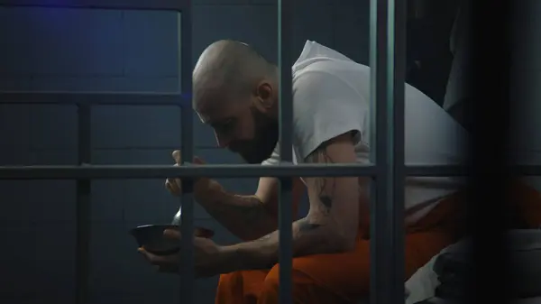 身穿橙色制服的男性囚犯坐在牢房的床上 吃铁碗里恶心的监狱食物 犯人因犯罪在狱中服刑 拘留中心或教养所 — 图库照片
