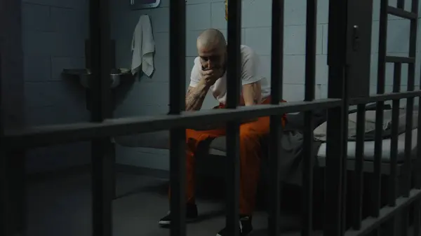 オレンジ色の制服の男性捕虜は刑務所のベッドに座り 鉄のボウルから嫌な刑務所の食べ物を食べようとします 刑務所では刑務所に収監されている 拘禁センターまたは矯正施設 — ストック写真