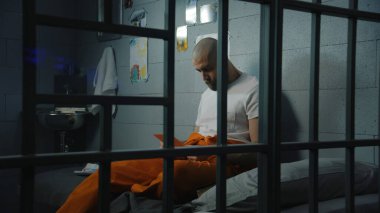 Turuncu üniformalı bir erkek mahkum hapishane hücresinde yatağında oturuyor ve aile ve çocukların fotoğraflarına bakıyor. Suçlu hapiste suçtan hapis cezasına çarptırıldı. Gözaltı merkezi ya da ıslah evi.
