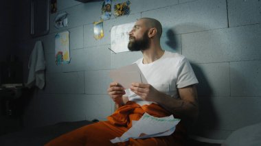 Üzgün erkek mahkum aile fotoğraflarına ve hapishane hücresindeki çocuk çizimlerine bakıyor. Suçlu hapiste suçtan hapis cezasına çarptırıldı. Gözaltı merkezi ya da ıslah evi. Portre.