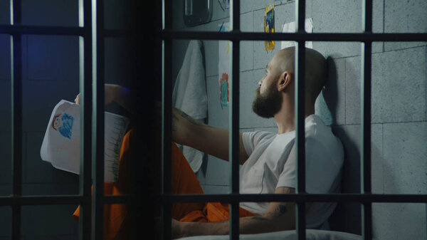 Depressed male prisoner holds drawings of child and looks at window sitting on bed in jail cell. Illegally convicted man serves imprisonment term in prison. Detention center or correctional facility.