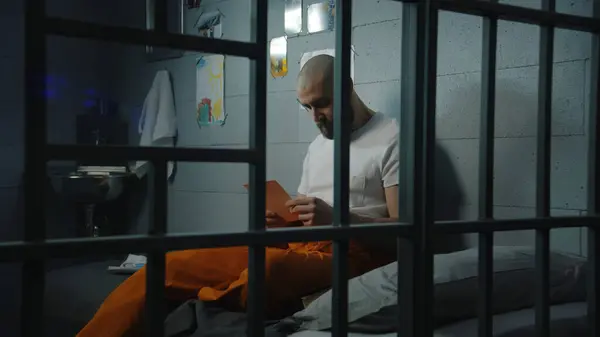 身穿橙色制服的男性囚犯坐在牢房的床上 看着家人和孩子的照片 罪犯因犯罪在狱中服刑 拘留中心或教养所 — 图库照片