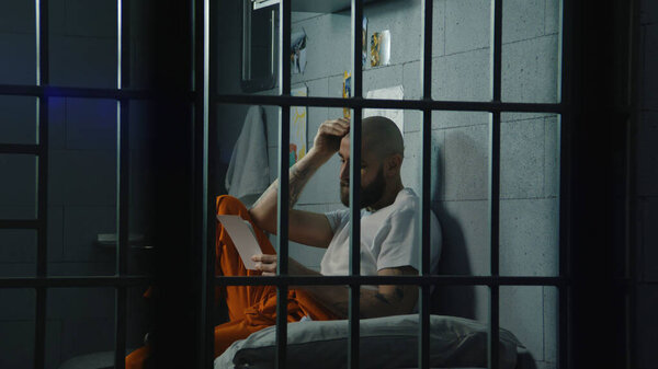 Upset male prisoner in uniform looks at pictures of family and kids sitting on bed in prison cell. Illegally convicted man serves imprisonment term in jail. Detention center or correctional facility.