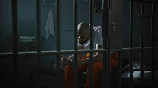 Criminal in orange uniform sits on prison cell bed. Prisoner serves imprisonment term for crime in jail. Gangster in detention center, correctional facility. Justice system. View through metal bars.