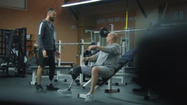 Modern spor salonunda halterli protez bacak egzersizleri yapan atletik bir adam ve kişisel fitness koçu. Yapay bacaklı bir atlet spor ekipmanları kullanarak ağırlık çalışması yapıyor. Fiziksel rehabilitasyon