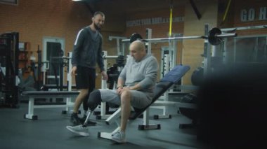 Modern spor salonunda halterli protez bacaklı ve kişisel spor eğitmeni olan motive bir sporcu. Fiziksel engelli yetişkin sporcu profesyonel spor malzemelerini kullanarak egzersiz yapıyor..