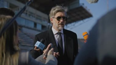 TV haberleri için basın toplantısında özel yatırımcıyla röportaj. Organizasyon temsilcisi basın sorularını yanıtlıyor ve bir futbol stadyumunun dışında röportaj veriyor. Gazeteciler basın kampanyasında kalabalıklar.